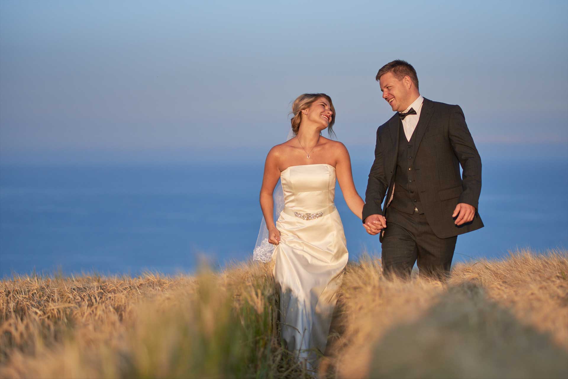 Wedding couple walk hand in hand in knee length grass against the backdrop of a big clear blue sky. They stare lovingly into each others eyes and are smiling.