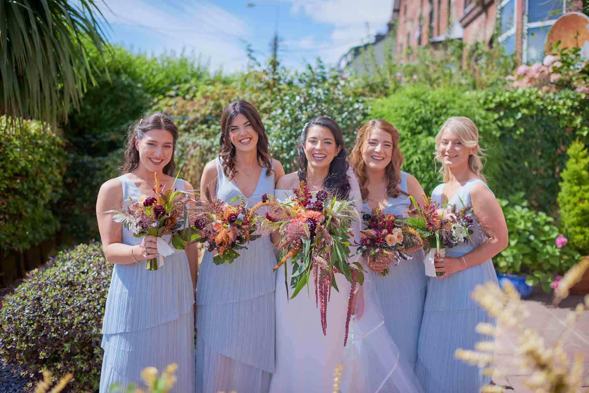 Bride lined up outsider her house in Cork city suburb with her 4 bridesmaids. Each hold a colourful wedding bouquet and dressed in a blue dress. The bride is smiling in her plain and elegant white dress