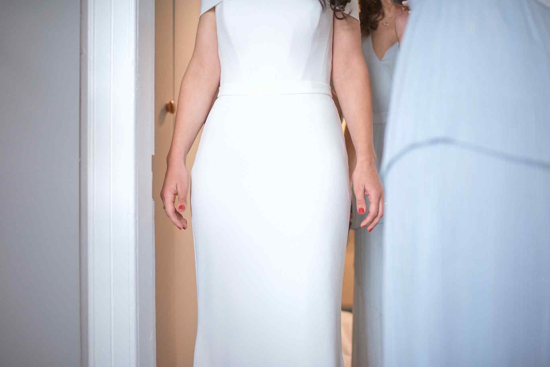 Bride with her arms by her side in her fitted white wedding dress. Her red painted fingernails stand out with the white of the dress.