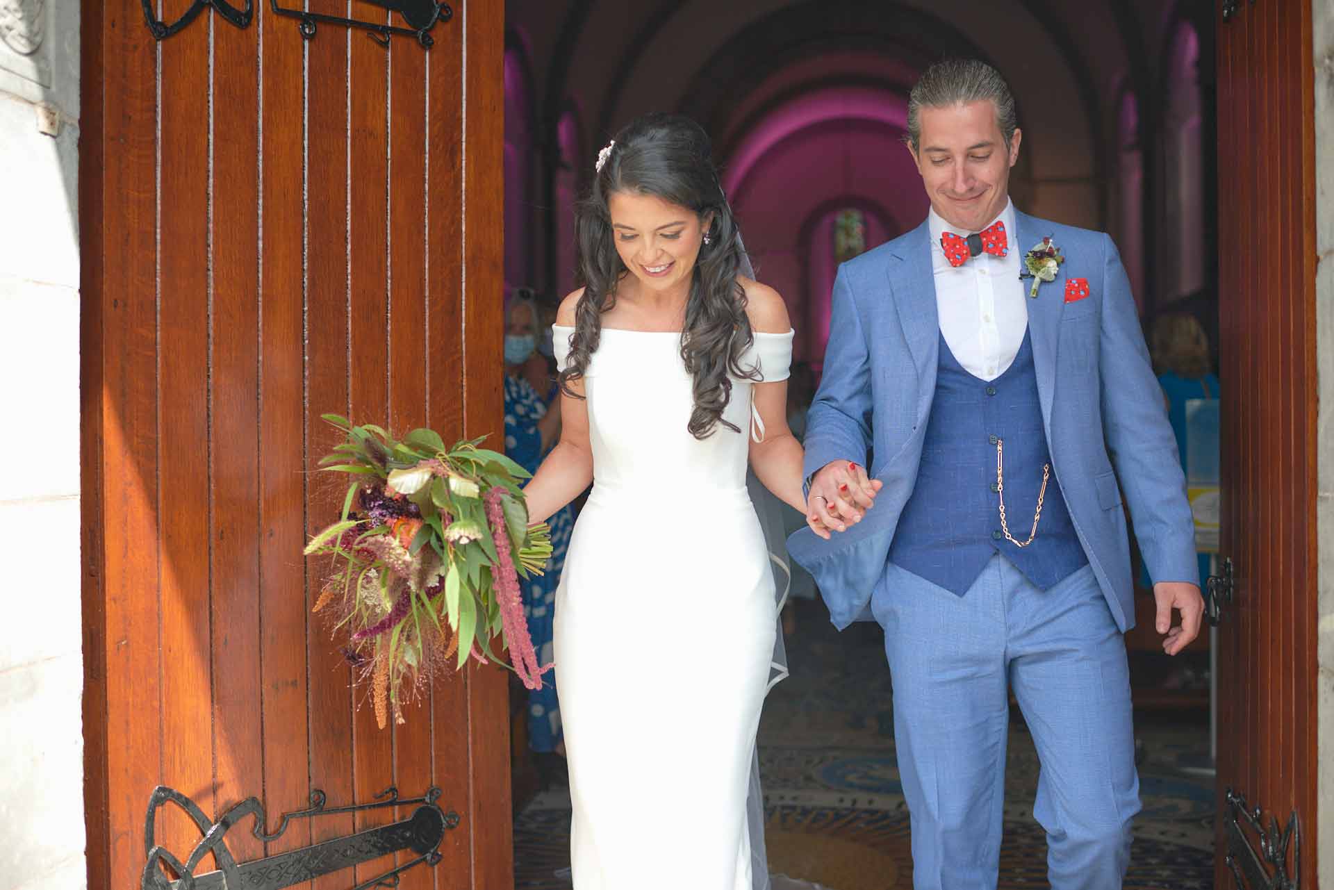 Bride and groom lock hand in hand leaving Honan chapel after getting married looking happy. Bride holds a big wedding bouquet of flowers in her right arm