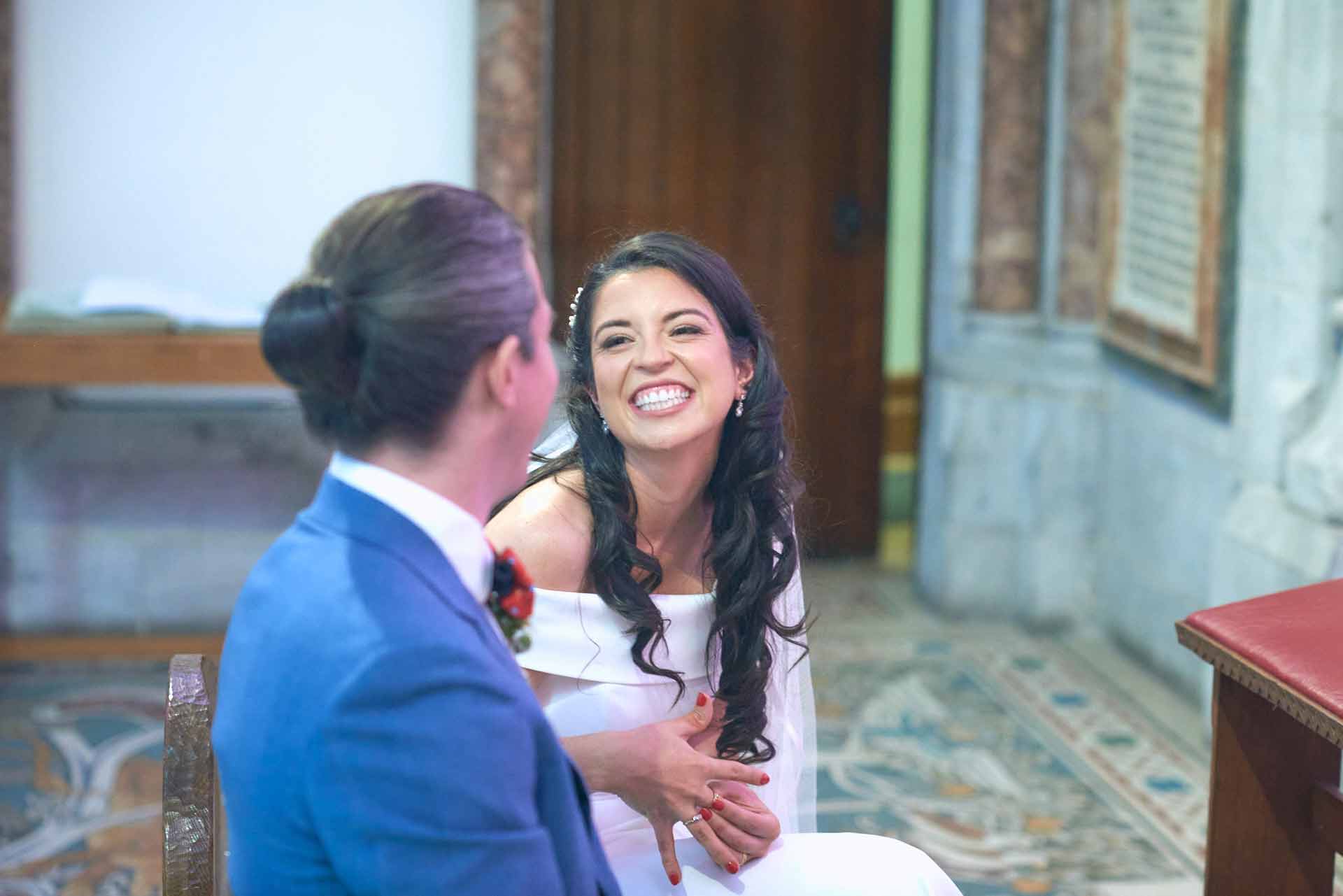 Ecstatic looking bride with a huge beaming smile feels her new wedding ring on her finger. They both sit on chairs close to the alter in the chapel.