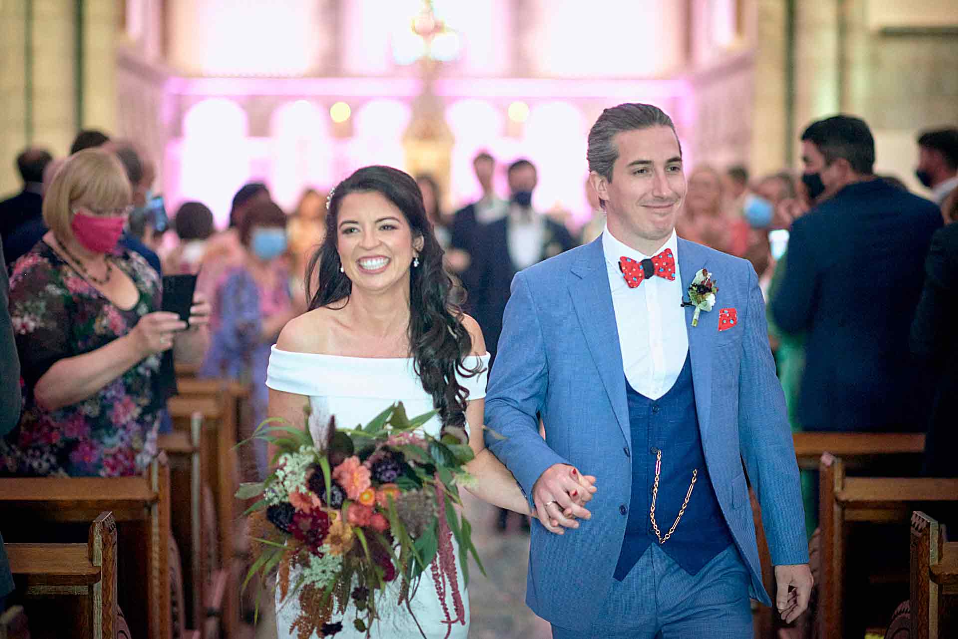 Bride and groom smiling arm in arm walk down the aisle. Bride in white dress is carrying wedding bouquet. Groom has a blue suit