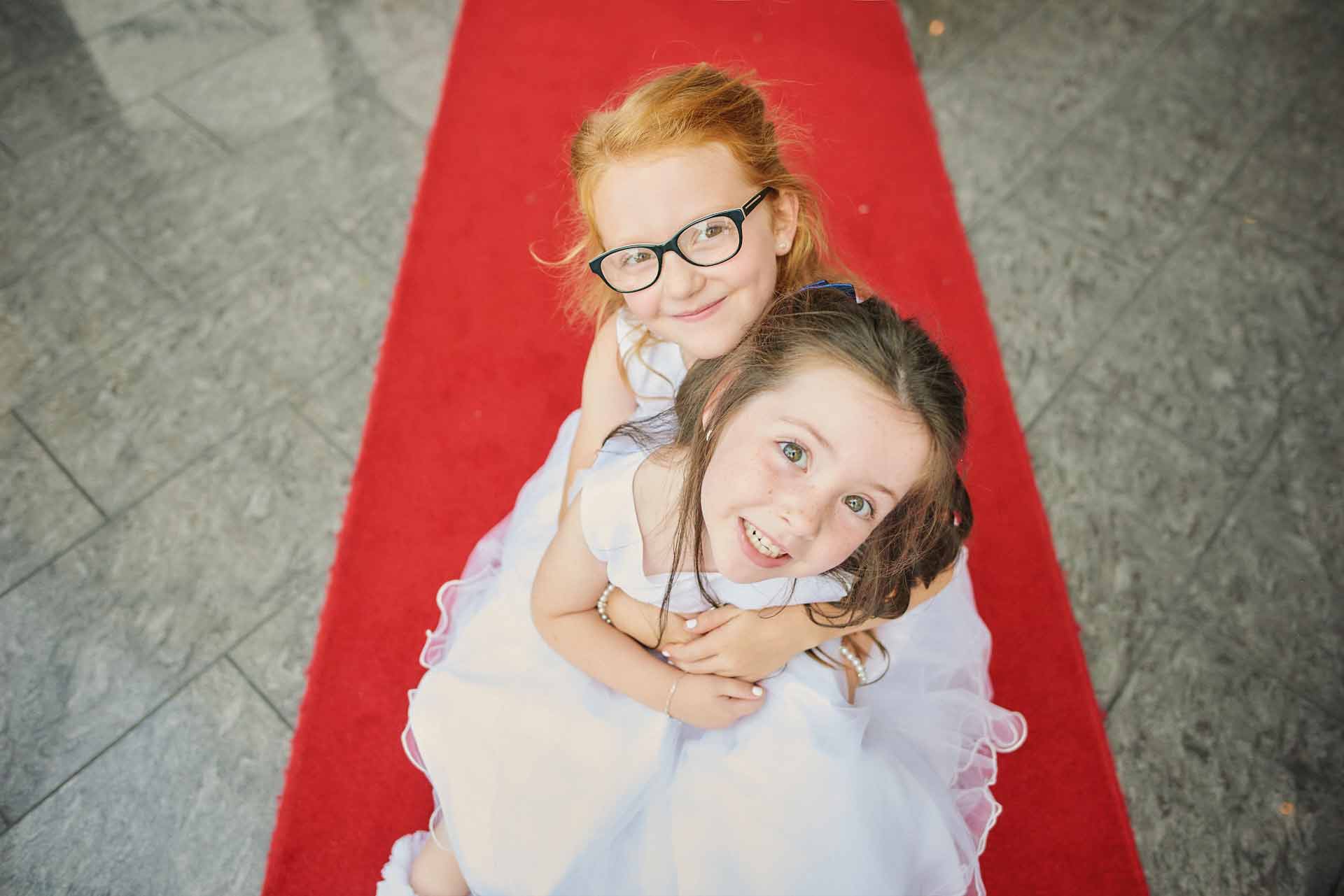 Two young flower-girls in their white wedding dresses pose happily together on a red carpet