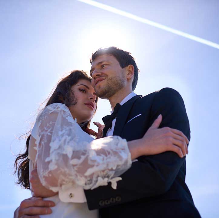 a low angle shot of the bride and groom holding each other close. The trail of an aeroplane leaves a white streak in the sky