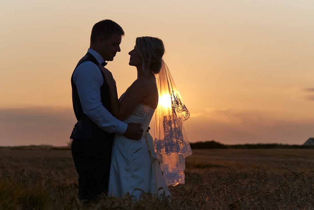 Silhouette of a bride and groom as they stand in a field watching the setting of the sun on a beautiful clear day.