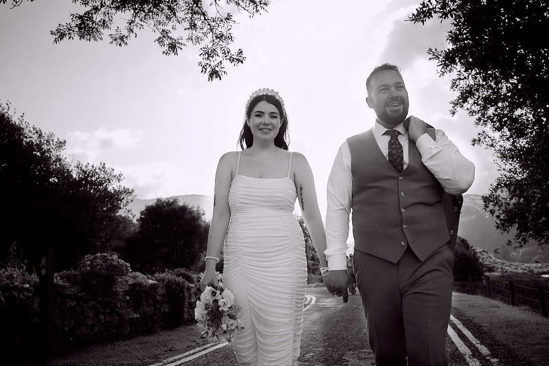 Happy bride and groom walking arm in arm down an empty road with trees in the background